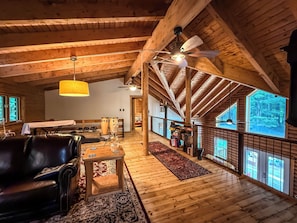 Een ruime kamer met een hoge plafond, houten balken, een plafondventilator en een groot raam met uitzicht op het buitengebied.