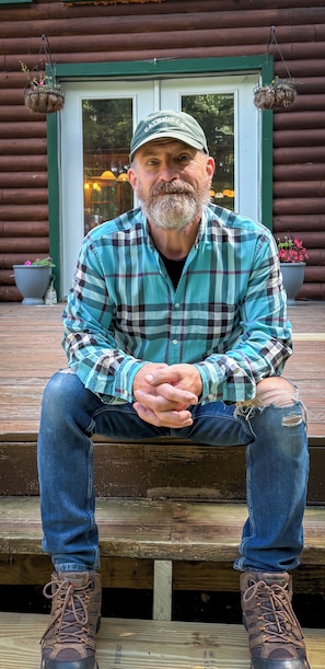 A man sitting on a wooden bench outside a log cabin with a green door and windows. - A Cozy Four Bedroom Log Home in the Heart of the Catskills (Saugerties)