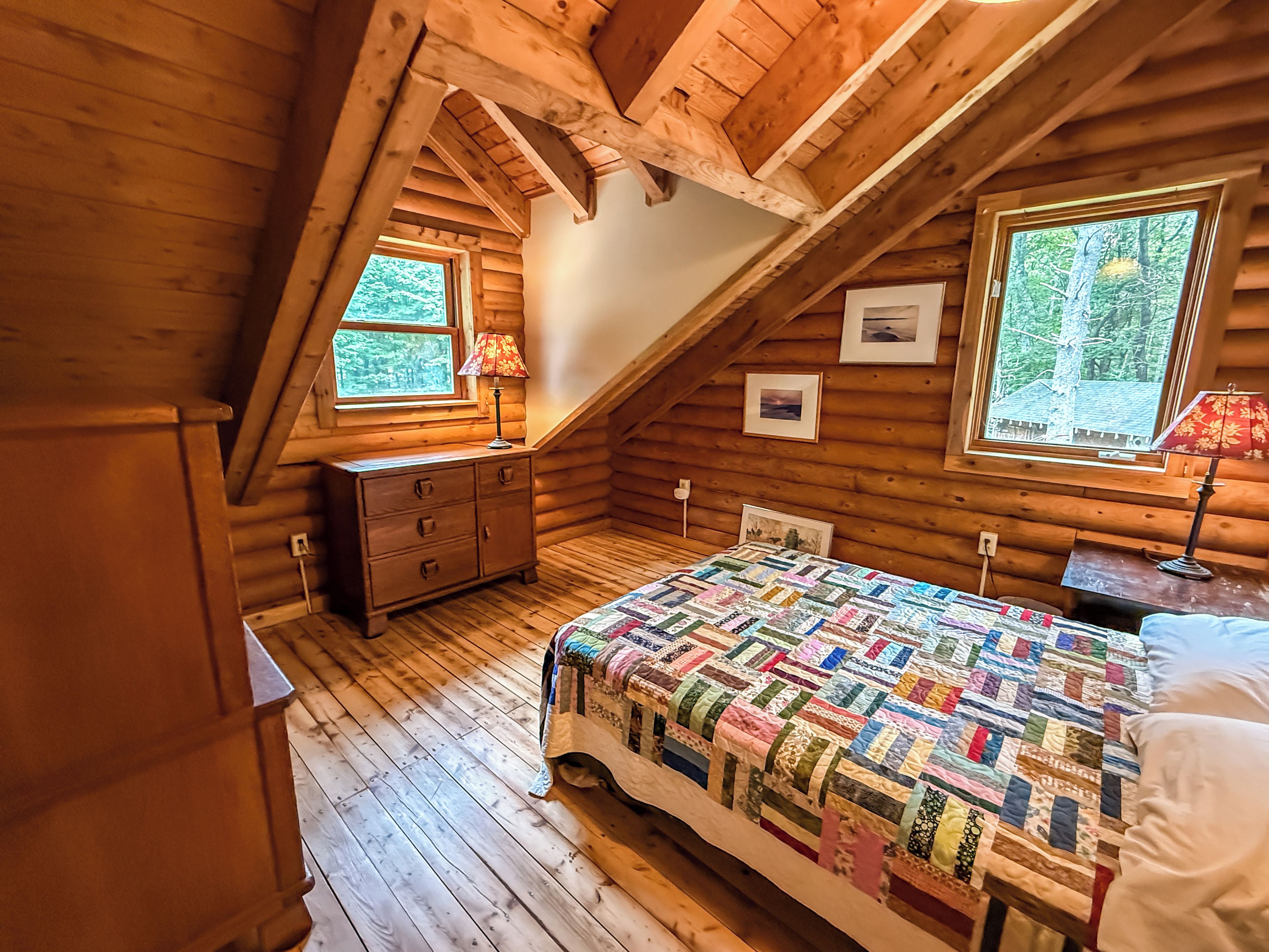 A log cabin bedroom with a wooden floor, a bed with a colorful quilt, a dresser, and two windows with lamps.
