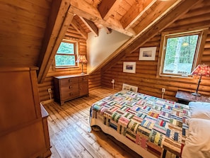 Une chambre dans une cabane en rondins, avec un plancher en bois, un lit recouvert d’une courtepointe aux couleurs vives, une commode et deux fenêtres avec des lampes.