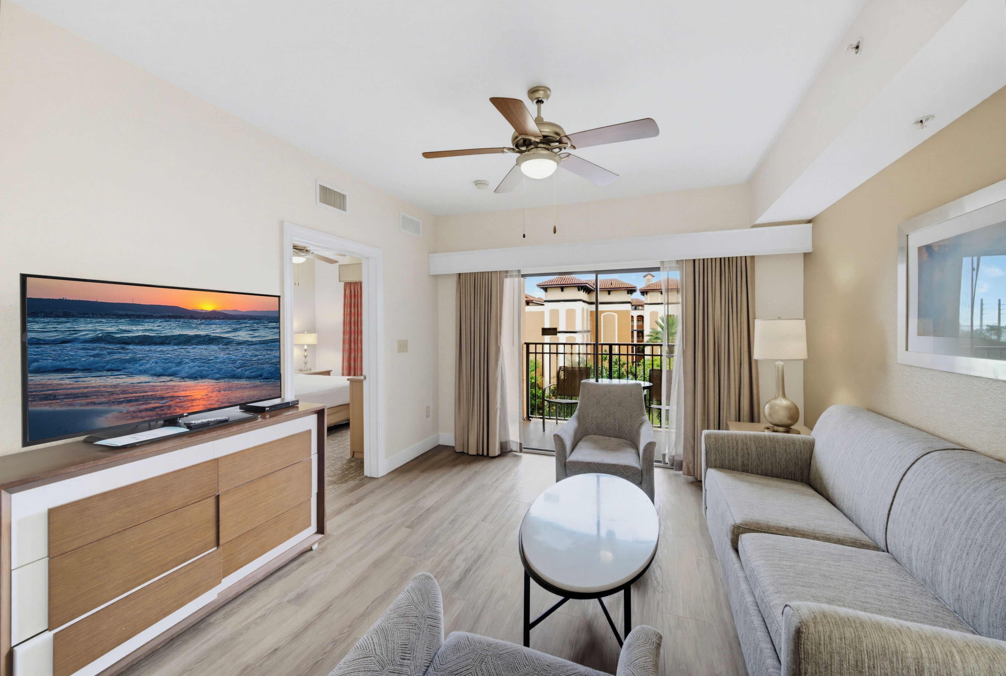 Ein Hotelzimmer mit Fernseher, Deckenventilator, Sofa, Couchtisch und einem Balkon mit Vorhängen.