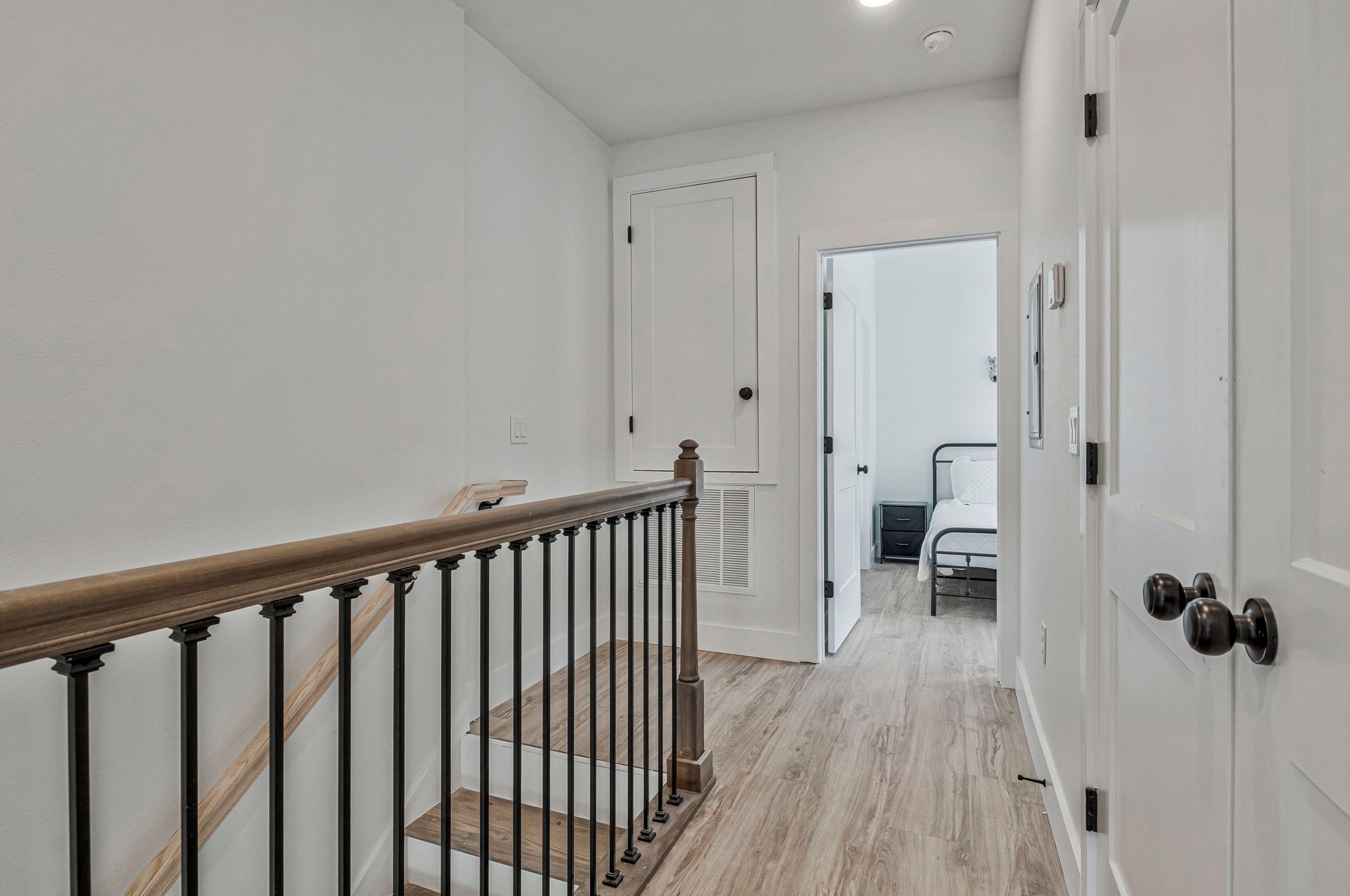 A staircase with a wooden handrail and black balusters leading to a bedroom with a bed and a nightstand.