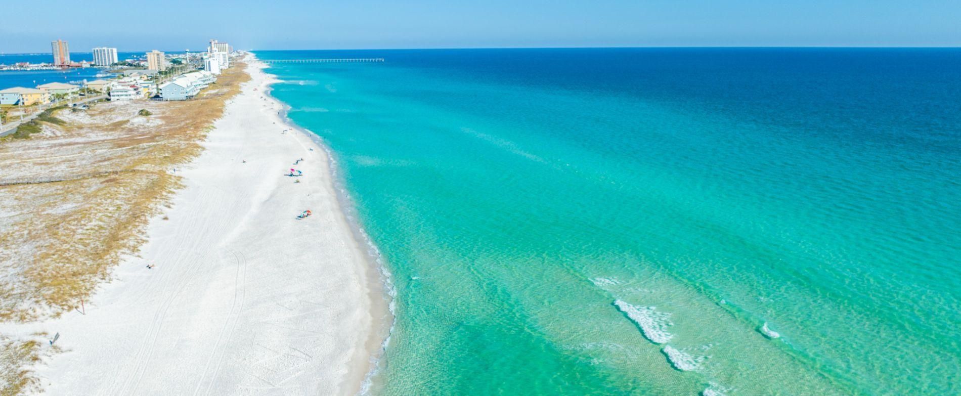 Una playa costera con vista despejada al océano, un muelle a lo lejos y edificios a lo largo de la costa.