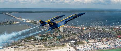 A jet performing a low-altitude maneuver over a coastal area with a beach, pier, and marina.