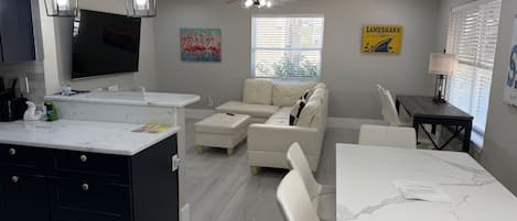 A modern kitchen with a central island, a dining area with a marble table, and a living space with a sofa and a ceiling fan.