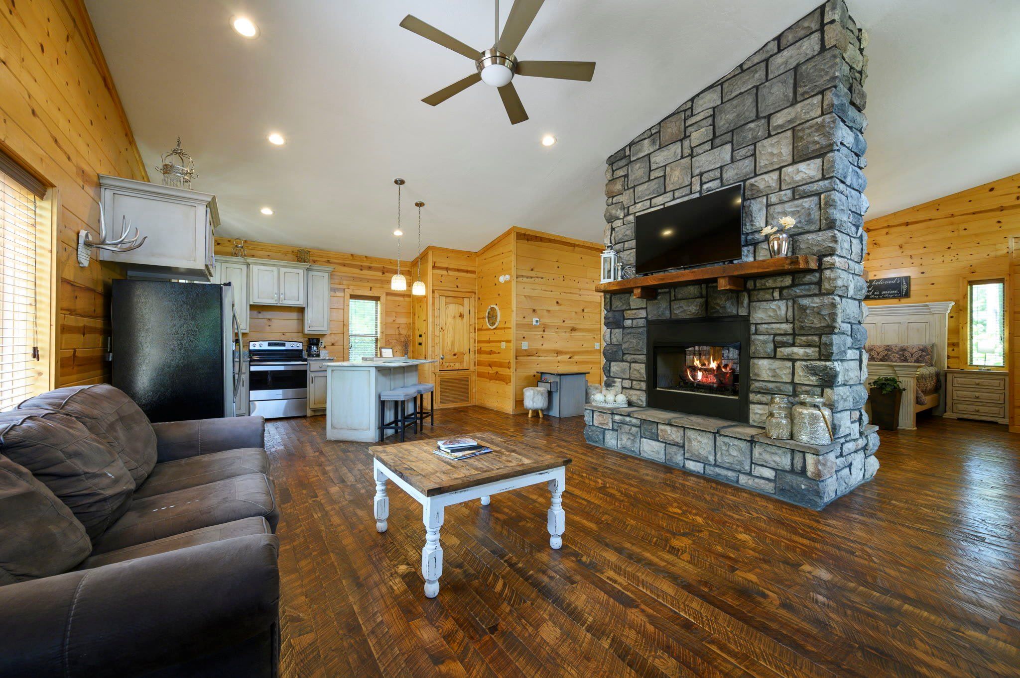 A log cabin interior with a stone fireplace, wooden floors, a ceiling fan, and a kitchen area.