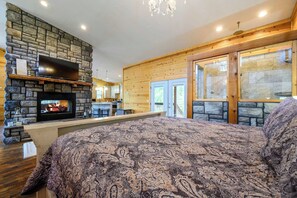 A cozy bedroom with a fireplace, a large bed, and a view of the kitchen and outdoor area. - Song of Songs - Private Hot Tub (Broken Bow)