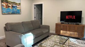 A living room with a gray sofa, a television on a wooden stand, and a painting on the wall. - The Land and Sea at Cape Neddick / A modern 1-bedroom apartment with AC (York)