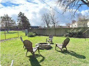 A backyard with a fire pit, three Adirondack chairs, a chain-link fence, and a building in the background.