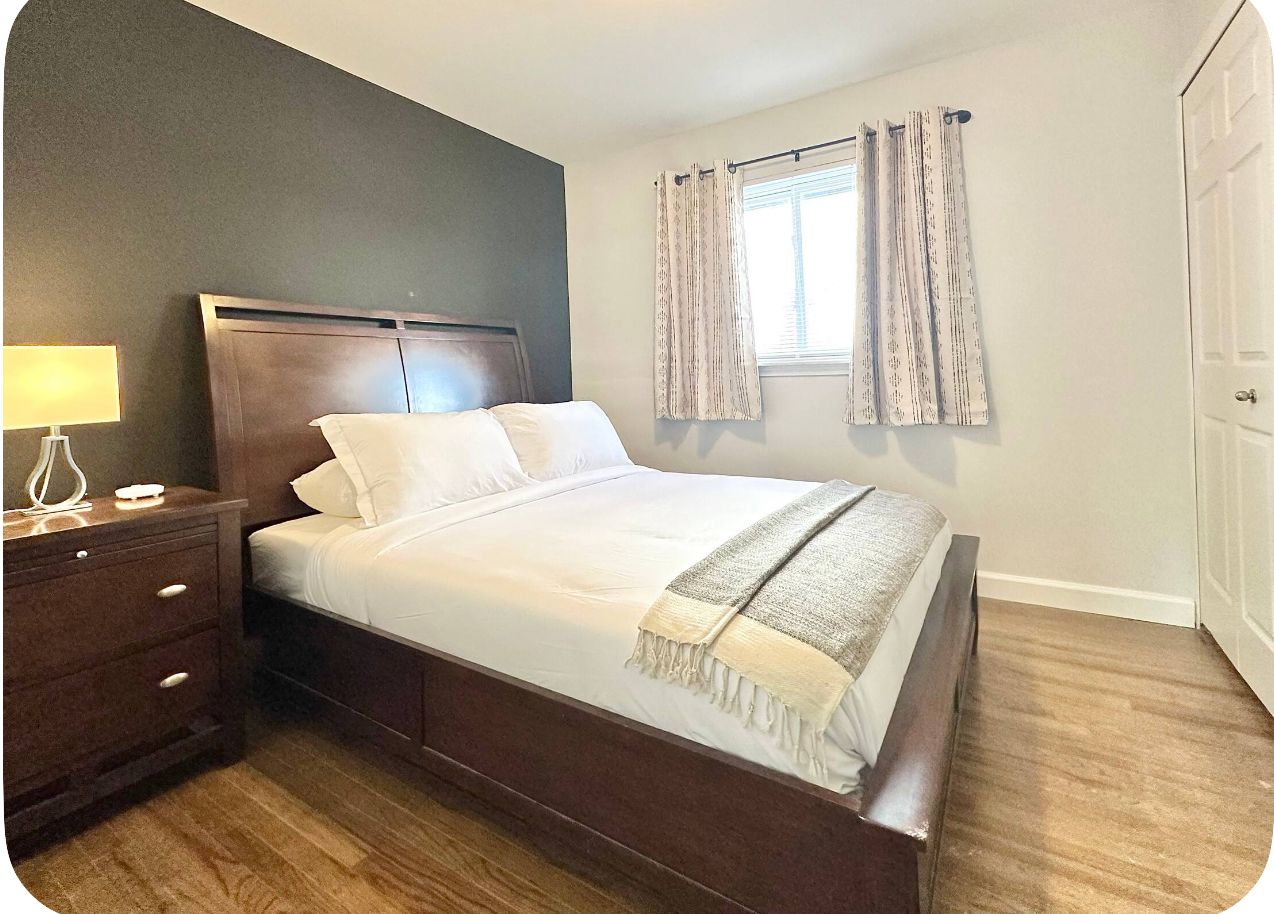 A bedroom with a wooden bed, a bedside table with a lamp, and a window with curtains.