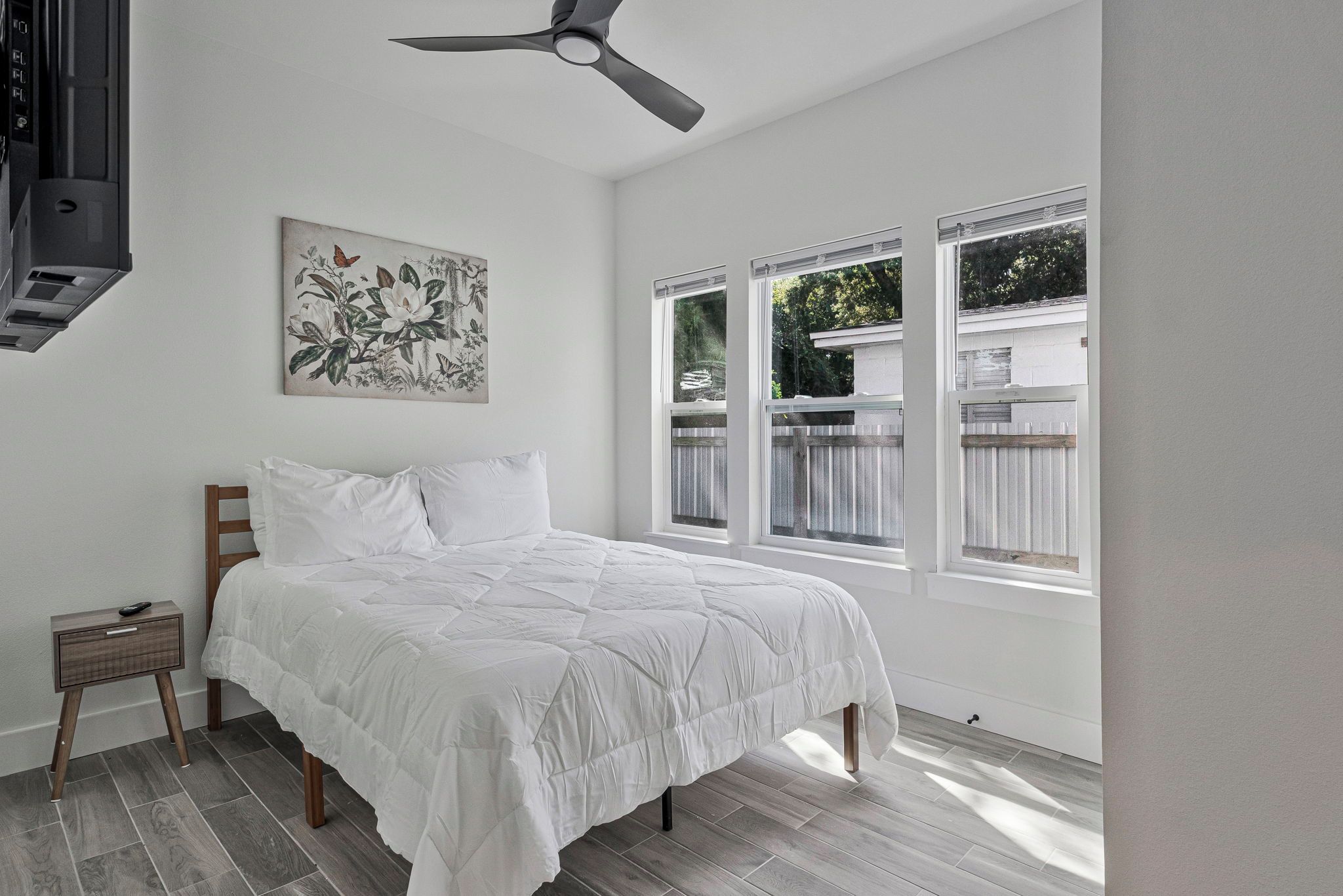 Une chambre à coucher avec un lit, une table de chevet, un ventilateur de plafond et une fenêtre donnant sur une clôture.