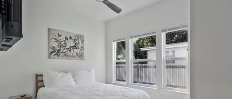 A bedroom with a bed, a nightstand, a ceiling fan, and a window with a view of a fence.
