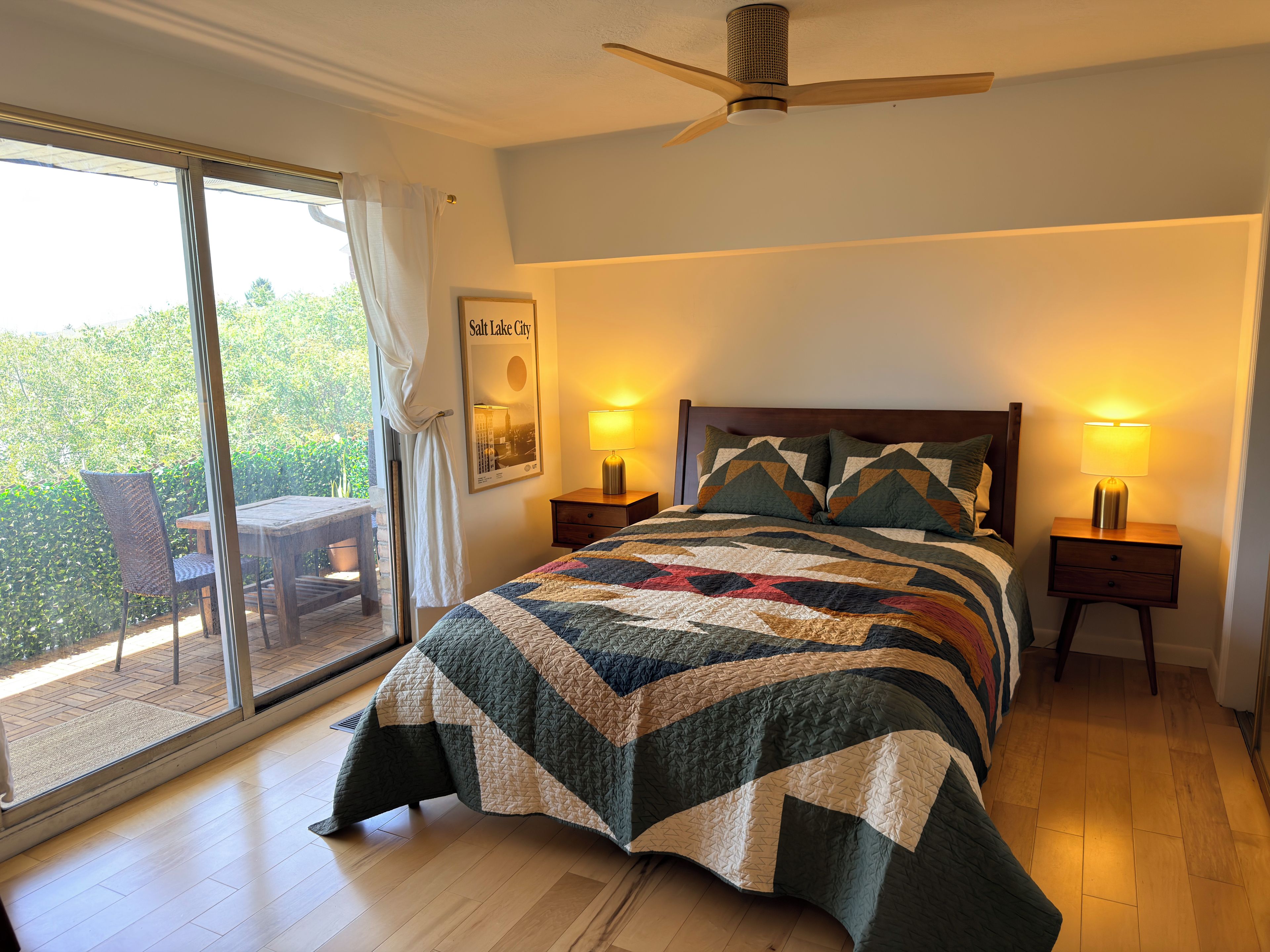 A bedroom with a large bed, two bedside lamps, a ceiling fan, a view of greenery outside, and a framed picture on the wall.