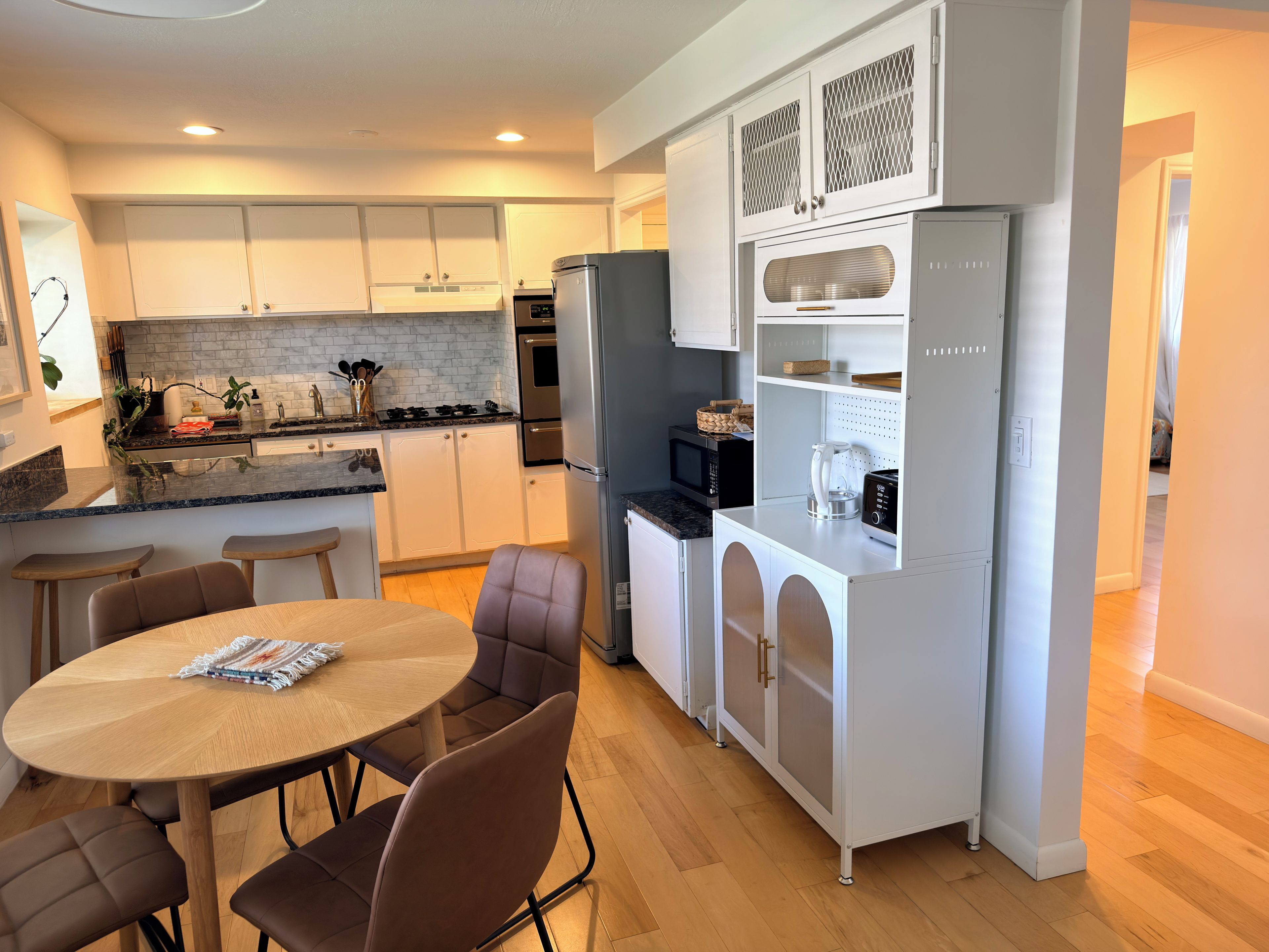 A modern kitchen with white cabinetry, a built-in oven, a microwave, and a refrigerator. There is a dining area with a round table and chairs.