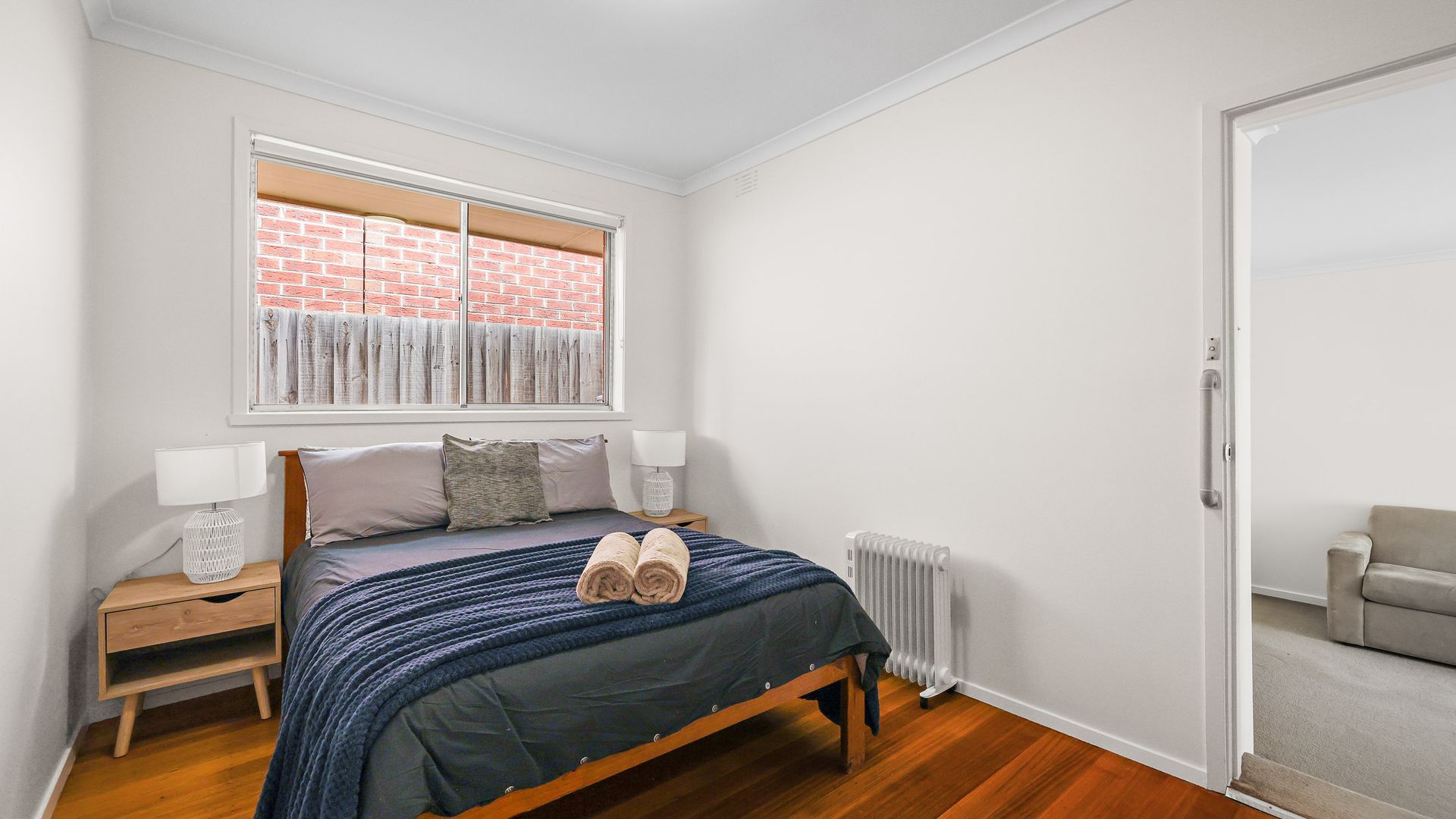A bedroom with a bed, bedside tables, a radiator, a window with a view of a brick wall, and a door leading to another room.