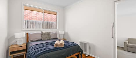 A bedroom with a bed, bedside tables, a radiator, a window with a view of a brick wall, and a door leading to another room.