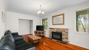 A living room with a black leather sofa, a wooden coffee table, a flat-screen TV on a wooden stand, a stone fireplace, and a large window with a view of trees. - Church Street Retreat (Cowes)