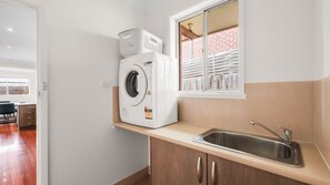 A compact kitchen with a built-in washing machine, a window with a view of a brick wall, and a stainless steel sink. - Church Street Retreat (Cowes)