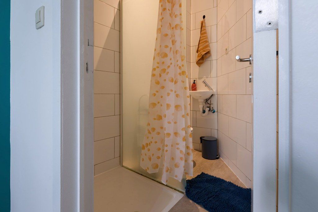 A bathroom with a shower, a towel rack, a sink, and a trash bin.