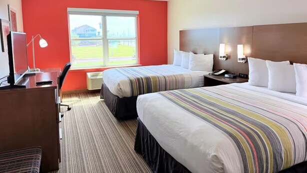 Ein Hotelzimmer mit zwei Betten, einem Schreibtisch, einem Stuhl, einer Schreibtischlampe und einem Fenster mit Blick auf ein Feld und Gebäude.