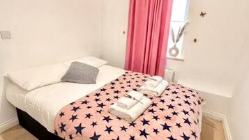 Ein Schlafzimmer mit einem Bett, einem Fenster mit Vorhängen, einer Wandleuchte und einem gerahmten Bild.