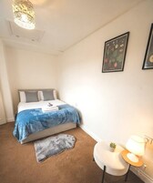 A bedroom with a bed, a bedside table with a lamp, a small rug, and a framed botanical illustration on the wall.