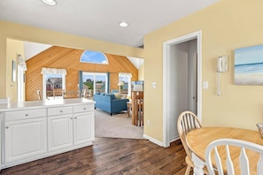 A kitchen with white cabinets, a wooden dining table, and a view of the backyard. - Beagle by the Sea | Hot Tub and Community Pool (Nags Head)