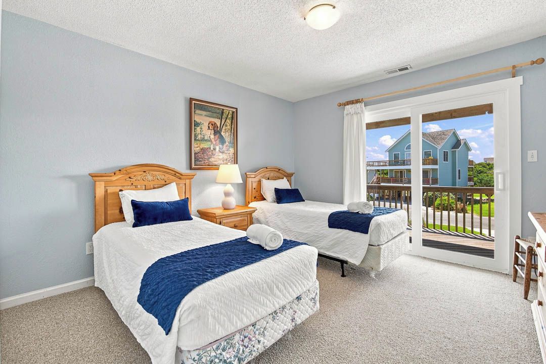 A bedroom with two beds, a nightstand, a lamp, a painting, and a sliding glass door leading to a balcony.