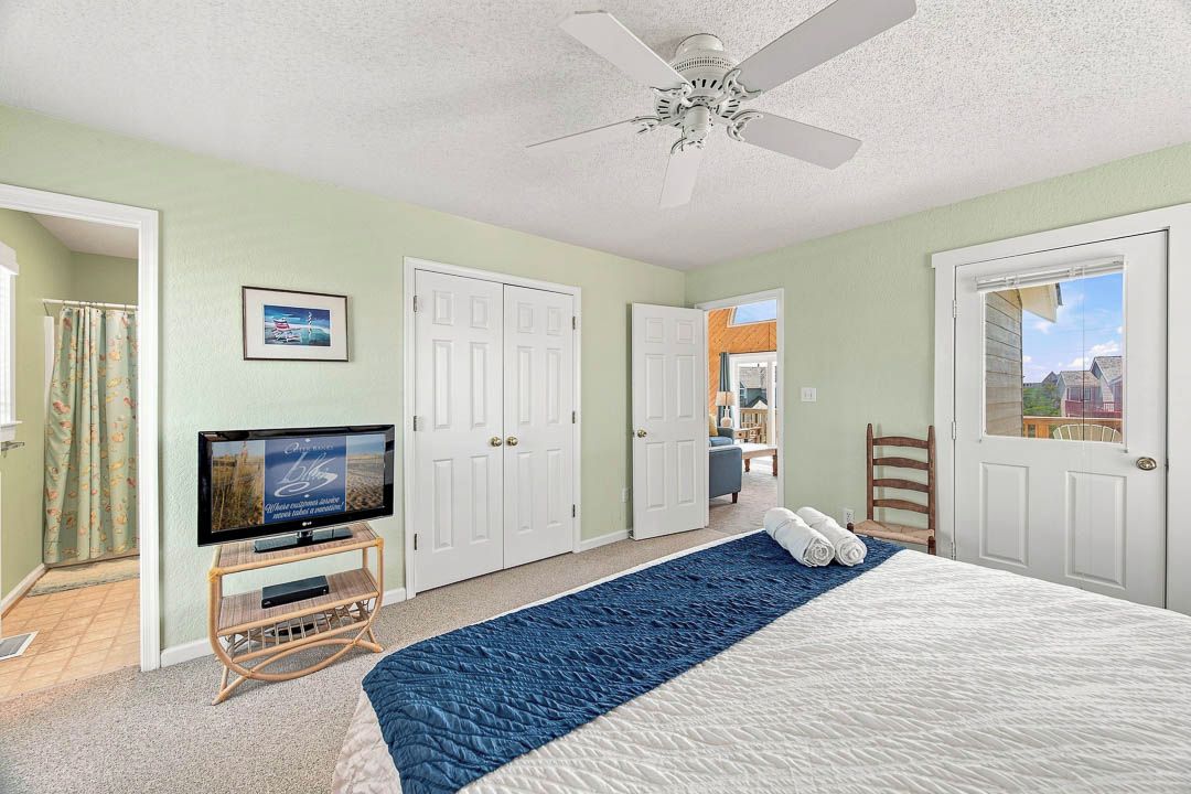 A bedroom with a bed, a television, a ceiling fan, and a view of the outside.