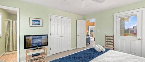 A bedroom with a bed, a television, a ceiling fan, and a view of the outside.