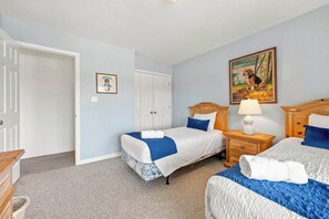 A bedroom with two beds, a nightstand with a lamp, a door, and a painting on the wall. - Beagle by the Sea | Hot Tub and Community Pool (Nags Head)