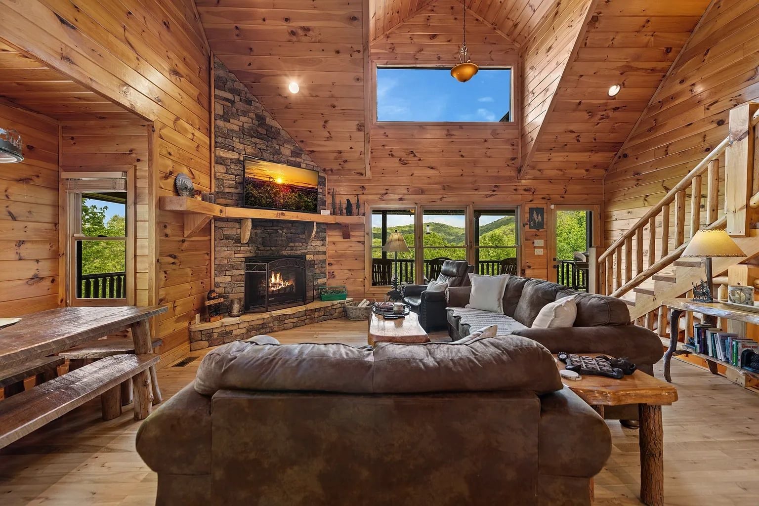 A log cabin interior with a stone fireplace, a sofa, a table, and a view of the outdoors.