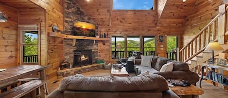 A log cabin interior with a stone fireplace, a sofa, a table, and a view of the outdoors.