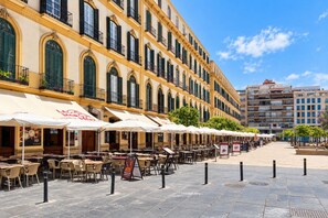 An outdoor cafe area with tables and chairs, a building with multiple floors and balconies, and a clear sky. - Guestready - Malaga Art and Comfort Apartments (Málaga)