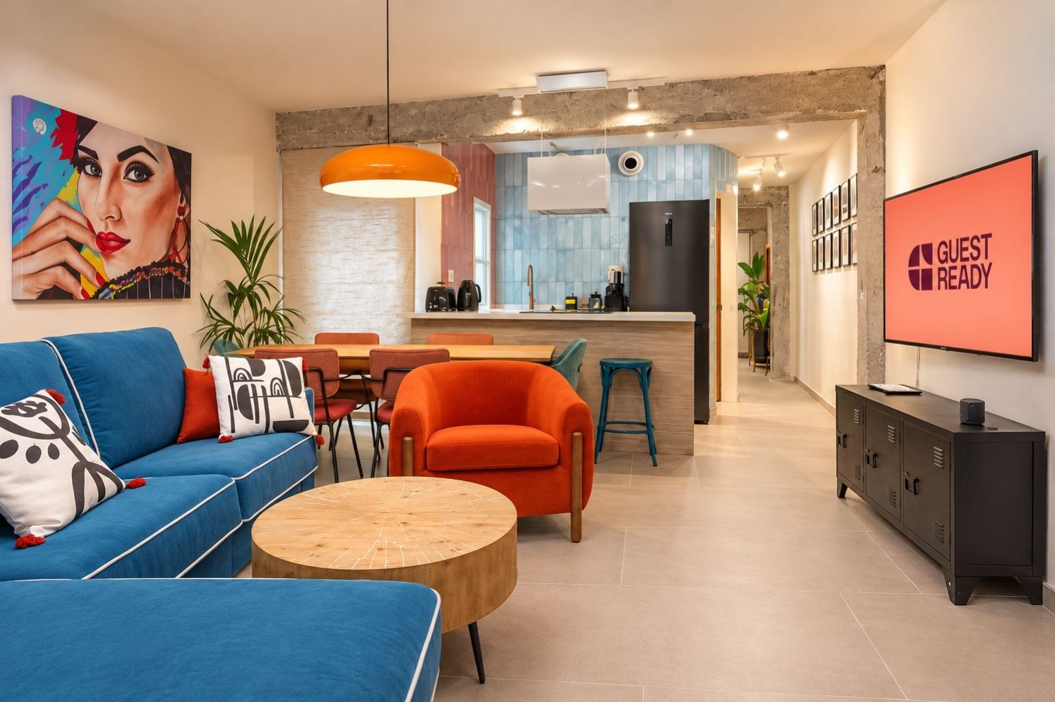 A modern living room with a blue sofa, orange armchair, and a round wooden coffee table. A kitchen area with a black refrigerator and a dining table is visible in the background.