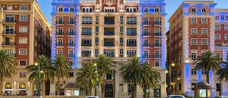 A multi-story hotel with a central dome and symmetrical facade, featuring balconies and palm trees in the foreground.