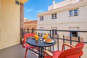 Un balcón con una mesa preparada para el desayuno, incluyendo croissants y zumo de naranja, y sillas rojas.