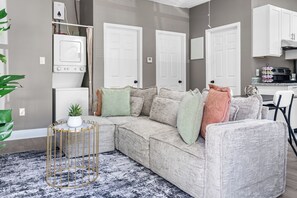 Ein modernes Wohnzimmer mit einer Eckcouch, einem runden Couchtisch mit Pflanze und einer Küche im Hintergrund.