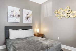 A bedroom with a bed, two wall-mounted abstract paintings, a bedside table with a lamp, and a decorative wall piece.