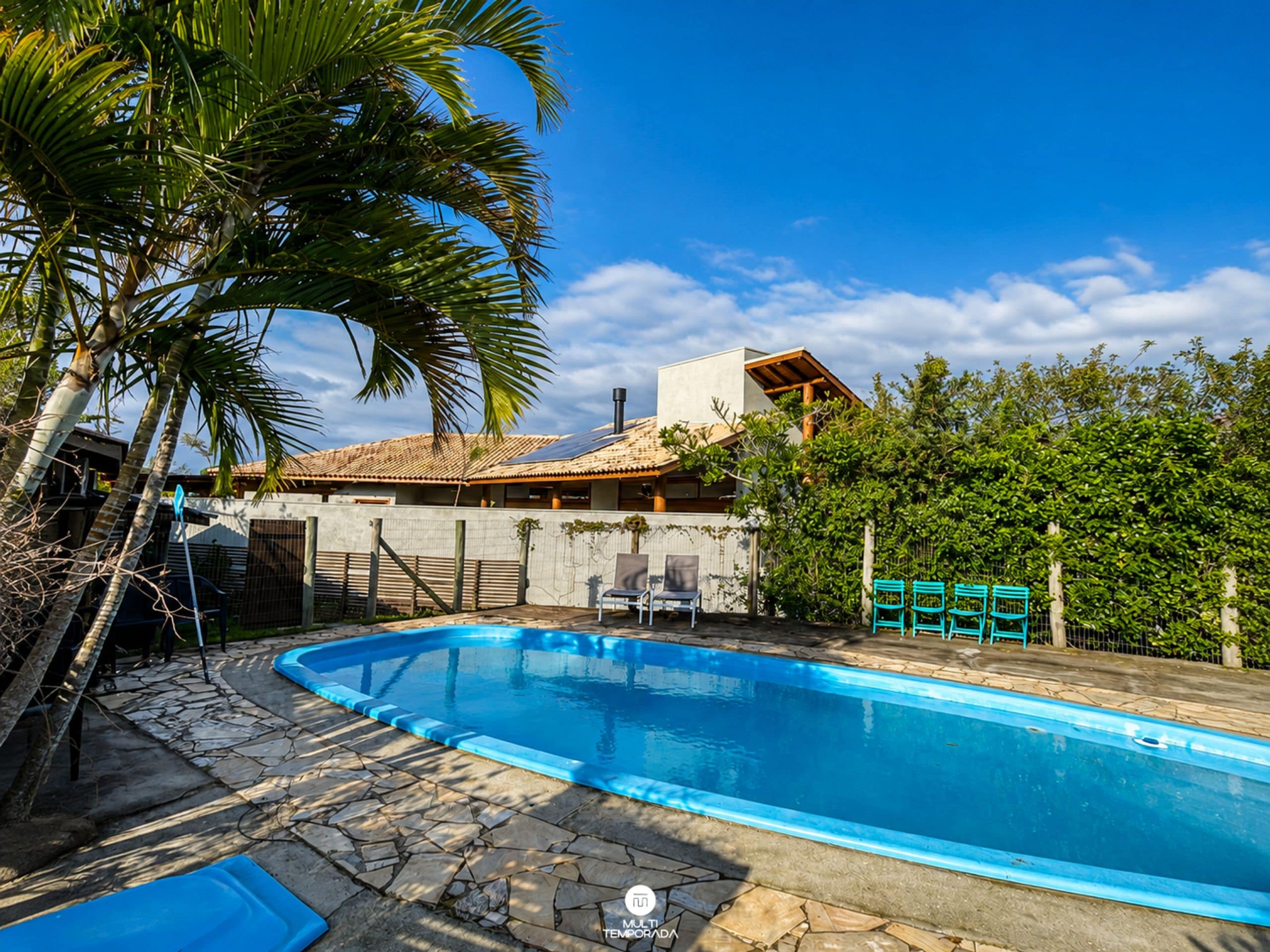 Una piscina con cubierta de piedra, rodeada de palmeras y una casa con techo de tejas.