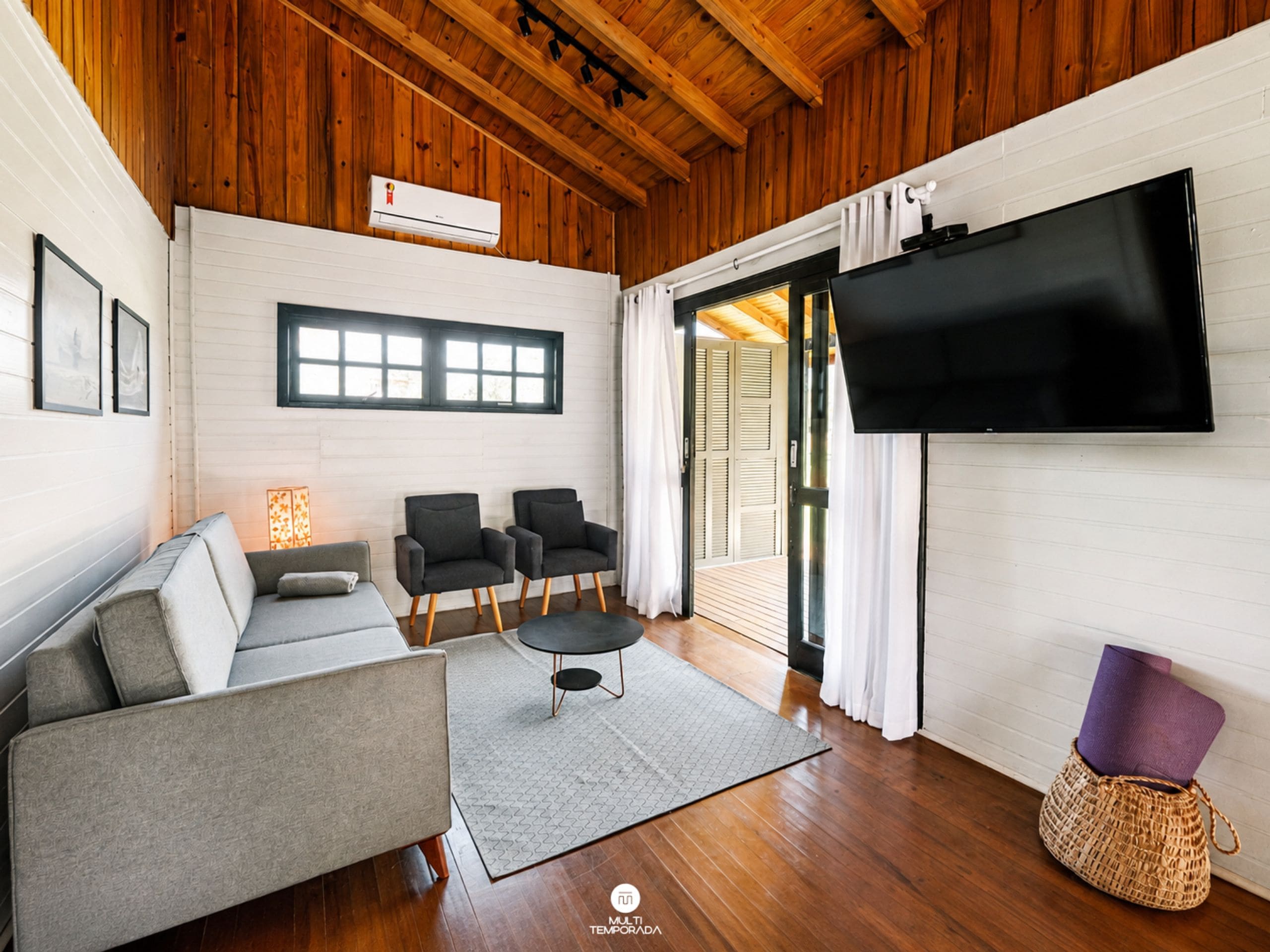 Uma sala de estar moderna com sofá, duas poltronas, mesa de centro, TV de tela plana fixada na parede e teto de madeira.