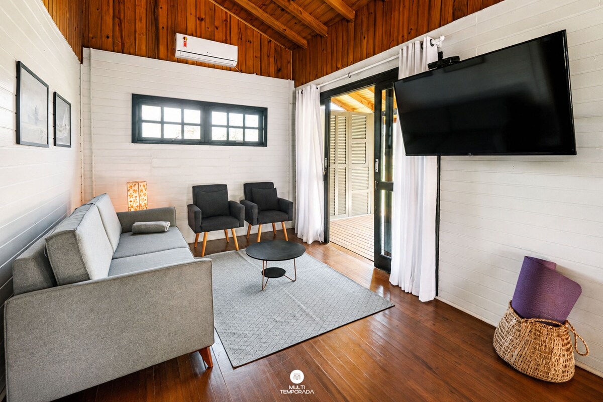 A modern living room with a sofa, two armchairs, a coffee table, a flat-screen TV mounted on the wall, and a wooden ceiling.