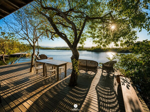 A wooden deck with a table and chairs, overlooking a calm lake with trees and a clear sky.