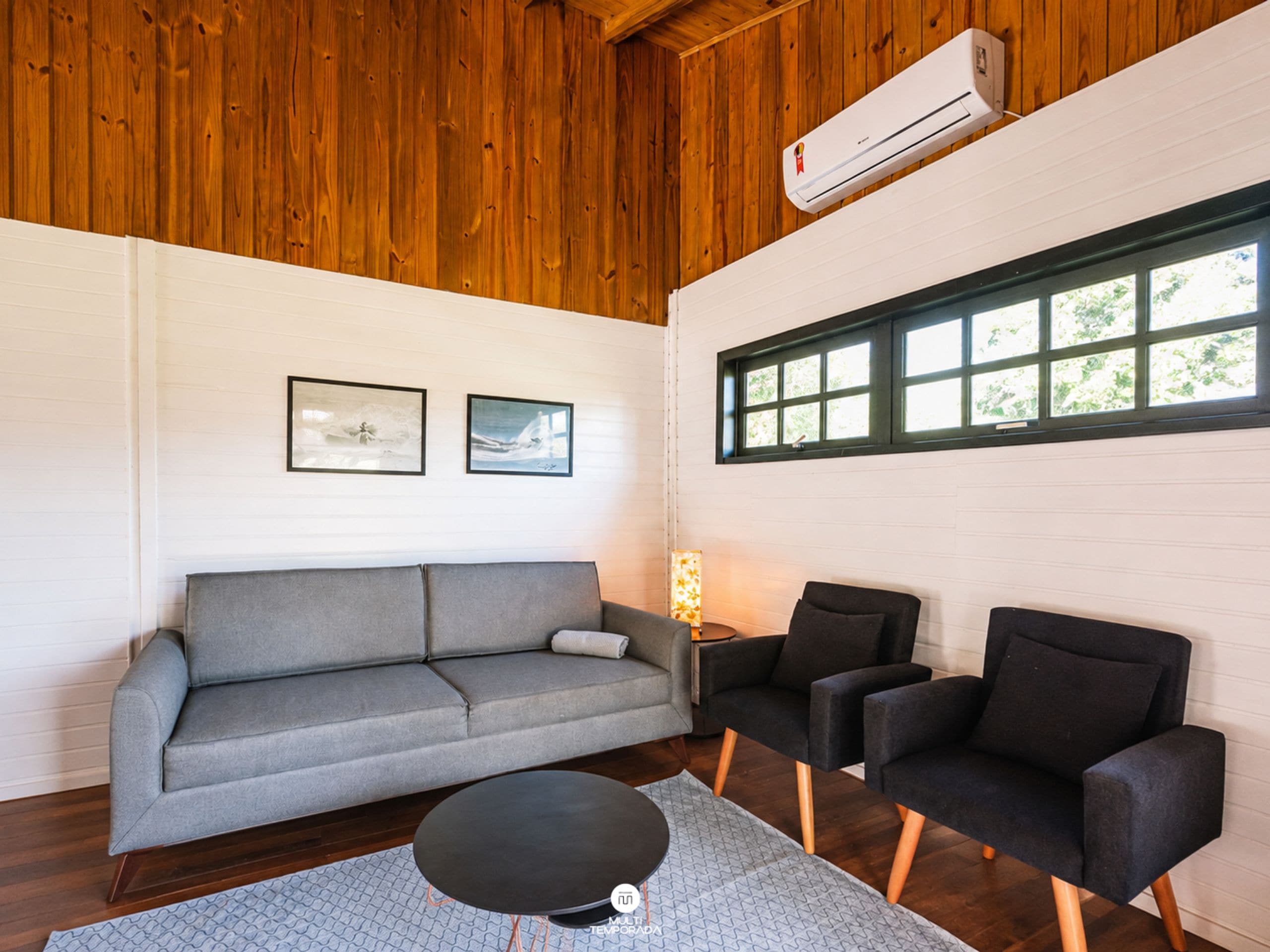 A room with a grey sofa, two black armchairs, a round coffee table, and a window with a view of greenery.