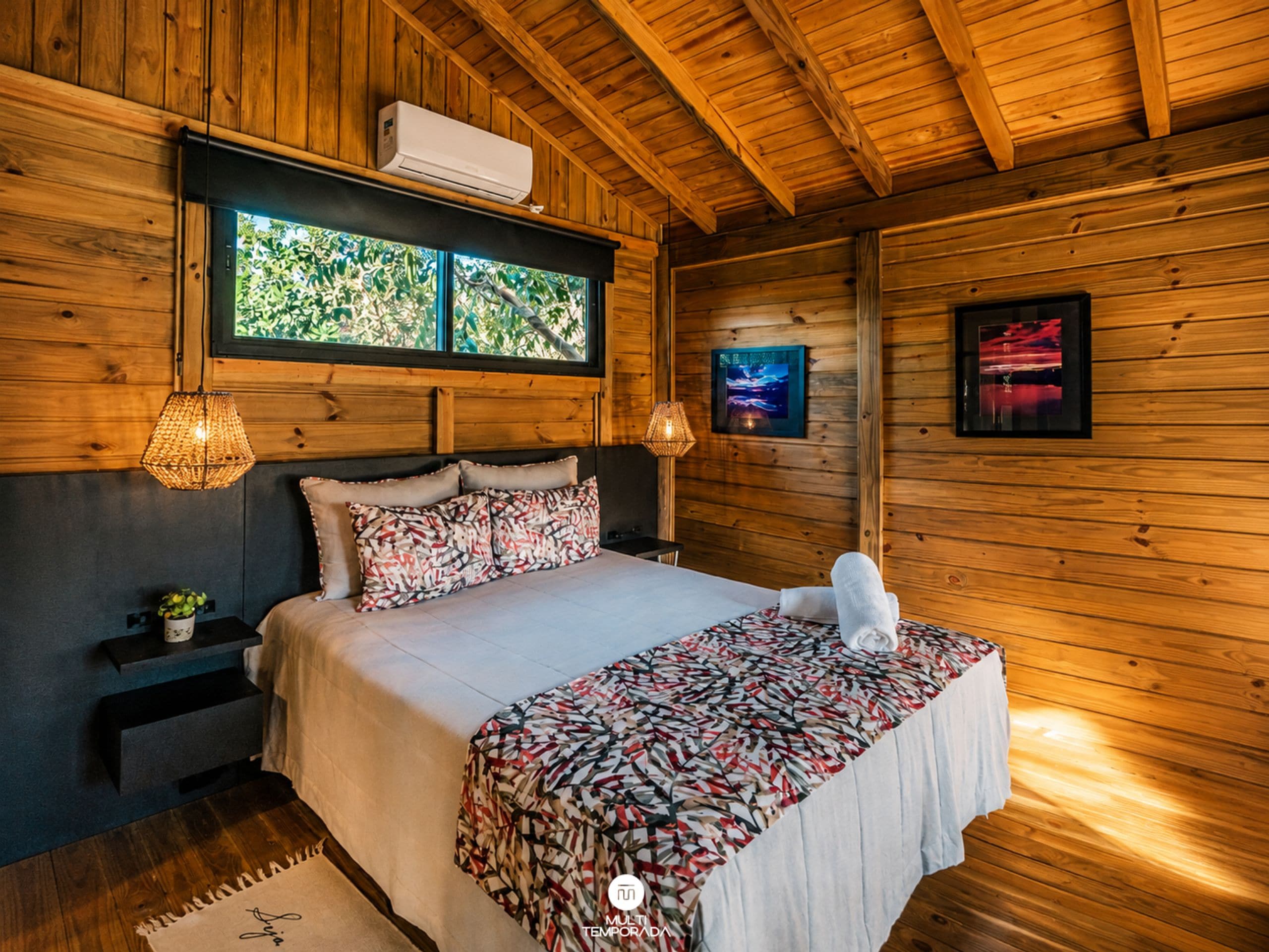 Quarto de cabana de madeira com cama, duas luminárias fixadas na parede, janela com vista e ar condicionado instalado.