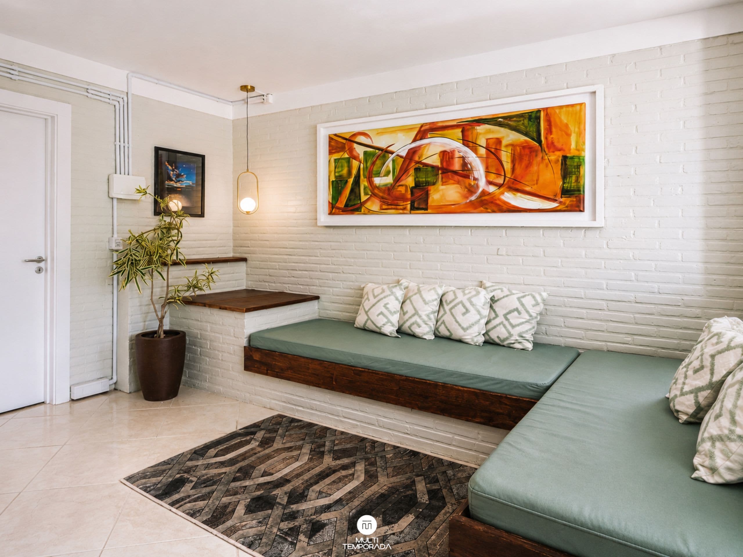 Uma sala de estar moderna com parede de tijolos brancos, um sofá verde com almofadas estampadas, um banco de madeira, uma planta em vaso e uma pintura abstrata grande.