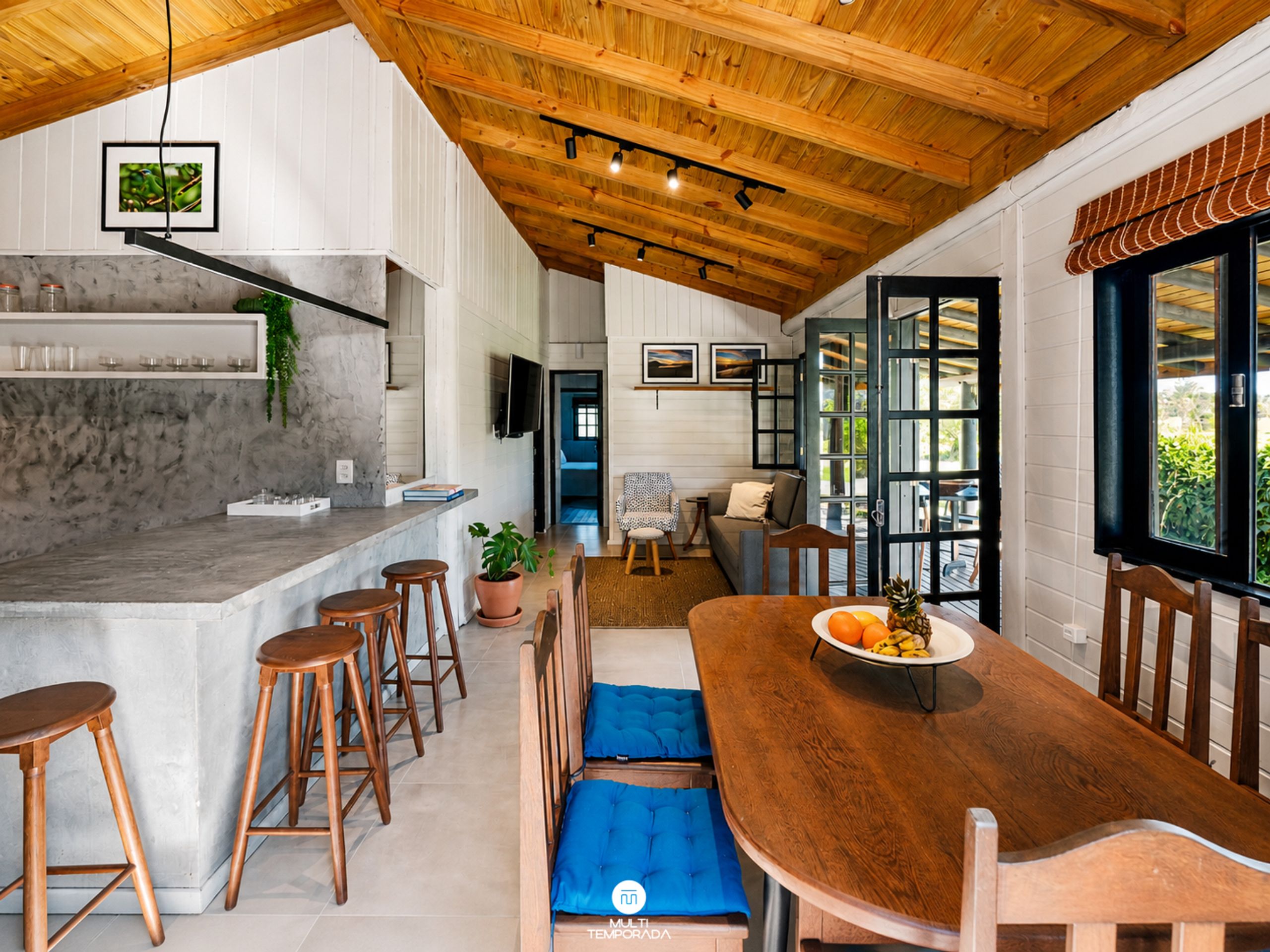 A modern kitchen with a marble countertop, wooden stools, and a dining area with a wooden table and blue cushions.