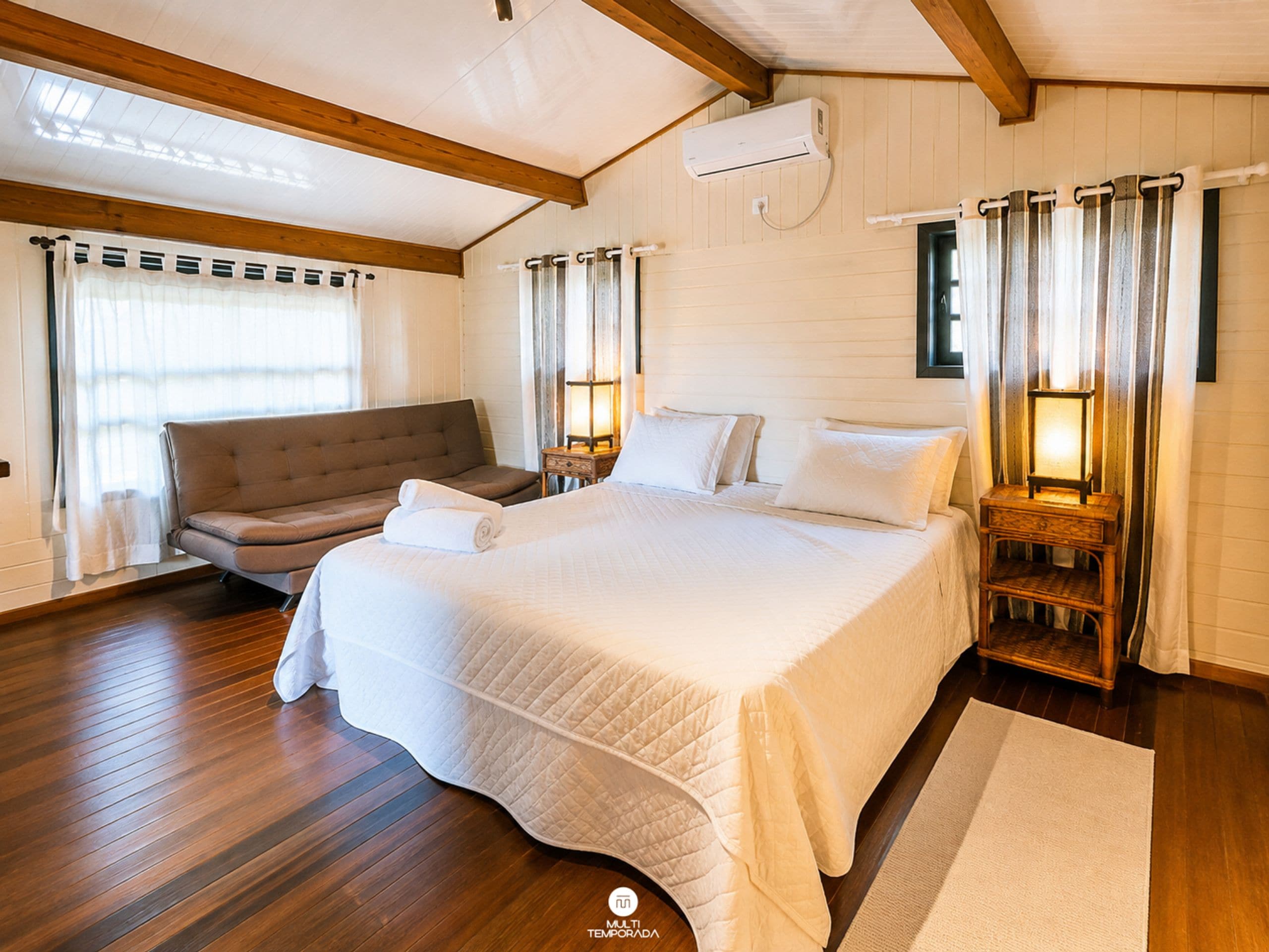 Quarto com uma cama grande, um sofá, uma mesinha de cabeceira com abajur e uma janela com cortinas.
