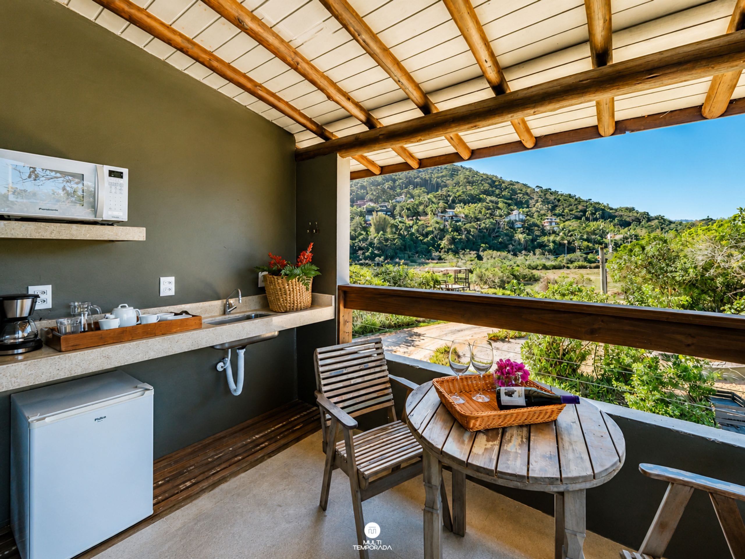 A balcony with a wooden table, a chair, a small refrigerator, a sink, and a view of a green landscape.