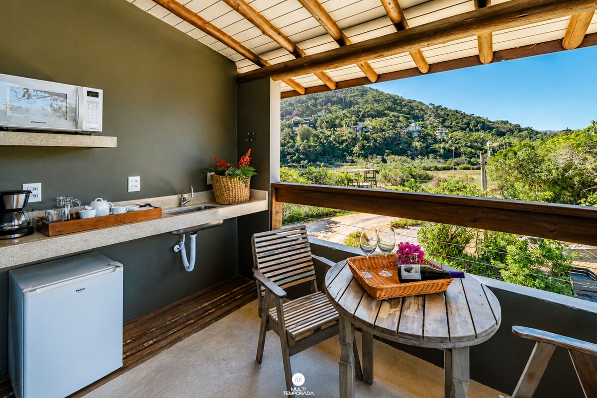 A balcony with a wooden table, a chair, a small refrigerator, a sink, and a view of a green landscape.